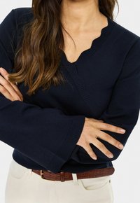 Femme les bras croisés, portant un pull bleu marine avec un col en V découpé et un pantalon crème avec une ceinture marron.