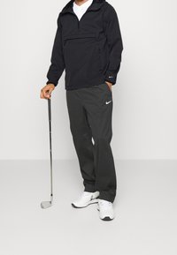 Mies mustassa Nike-verryttelypuvussa, joka pitää golfmailaa kädessään, valkoiset Nike-tossut jalassa, seisomassa yksinkertaisen valkoisen taustan edessä.