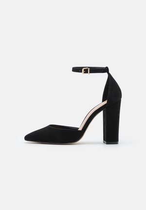 Sort ruskind højhælet sko med spids tå, ankelrem og blokhæl. Har en glat overflade og en detaljeret spænde i guldfarve.