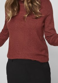 Femme aux cheveux blonds ondulés portant un pull en tricot couleur rouille et un pantalon noir, sur un fond gris uni.