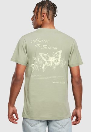 Lys grøn t-shirt med et stort hvidt sommerfugle- og blomsterdesign på ryggen, med teksten "Flutter & Bloom" i en elegant skrifttype.