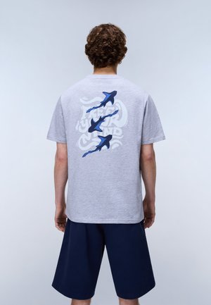 Person med krøllet hår står med ryggen til og har en lysegrå t-shirt på med tre blå hammerhajer og tekst, kombineret med marineblå shorts.