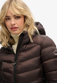 Chaqueta de abrigo marrón con cierre de cremallera, collar alto y capucha. Presenta un diseño acolchado, un parche de logo en el brazo izquierdo y una textura suave.