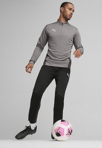 Grijs sportpullover met een ritskraag, gecombineerd met zwarte sportbroek en zwarte voetbalschoenen. Bevat een roze en witte voetbals.