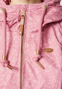 Rosa jacka med blommönster, metallisk dragkedja och snören. Har guldfärgade detaljer, huva och en slät textur.