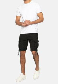 Homme portant un t-shirt blanc, un short cargo noir avec poches et des baskets blanches, se tenant contre un fond uni.
