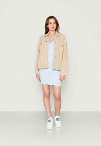 Jaqueta de ganga bege com bolsos frontais, vestido azul claro e sapatilhas brancas com manchas coloridas. Figura de pé sobre um fundo neutro.