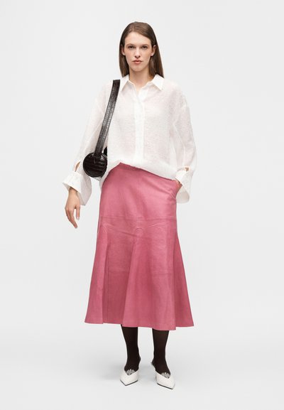 Λευκή, υφασμάτινη μπλούζα με φαρδιές μανσέτες, σε συνδυασμό με ροζ φούστα μήκους έως το γόνατο. Το μοντέλο φοράει μαύρες καλτσοδέτες και λευκά παπούτσια με διακοσμητικές λεπτομέρειες.