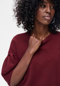 Bordowy, oversize sweter wykonany z miękkiego materiału, z prążkowanym dekoltem i luźnymi rękawami, z widocznymi detalami ramion i dłoni.