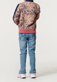 Bambino in piedi con le spalle rivolte verso la telecamera, indossando una giacca a motivo multicolore, jeans blu e sneaker rosa e bianche.