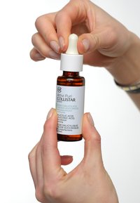 Amber glazen druppelfles met een witte dop; etiket bevat de productnaam en beschrijving in meerdere talen. Hand die de fles vasthoudt.