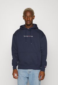 Sudadera azul marino con bolsillo frontal y capucha con cordón. Logo bordado de "Tommy Jeans" en blanco a lo largo del pecho. Textura suave.