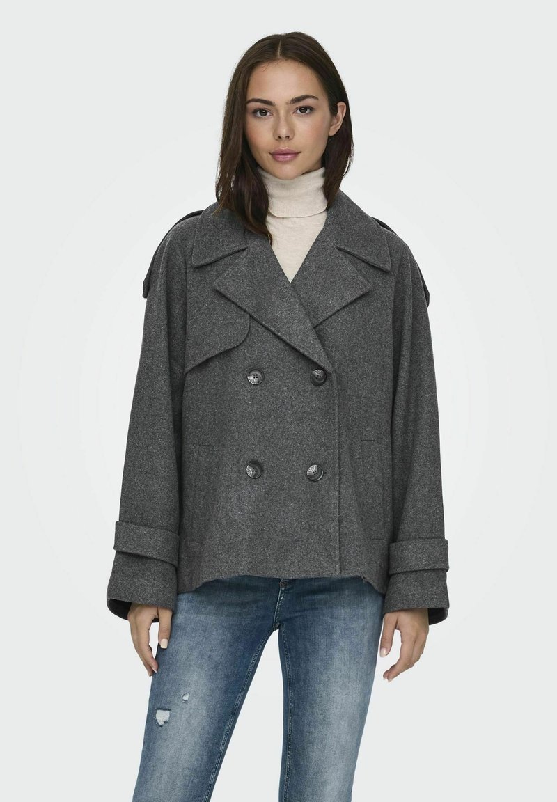 Veste en laine grise à double boutonnage avec grands revers, manches retroussées et deux poches avant. Portée par-dessus un col roulé beige et un jean bleu.