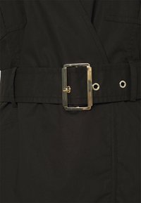 Czarny płaszcz z szerokim materiałowym paskiem z prostokątną złotą klamrą oraz srebrnymi oczkami. Materiał jest gładki o matowym wykończeniu.