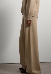 Pull en laine beige avec des poignets côtelés, associé à un pantalon en laine beige à jambes larges, surface texturée et fond uni et neutre.