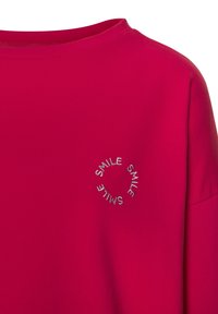 Camiseta de algodón roja con escote redondo y mangas cortas. Presenta un texto bordado en plata "SONRÍE" en un patrón circular en la parte frontal.