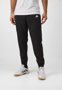 Black athletic pants with a slim fit, made of lightweight fabric. Features a white Adidas logo on the left thigh. Worn with white sneakers.