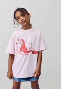 Tricou roz cu mâneci scurte, cu un design grafic roșu reprezentând o fată, accentuat de cuvinte, pe un fundal neutru. Fustă din denim vizibilă.