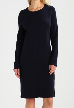 Femme portant une robe en maille bleu marine, manches longues, longueur genou, avec un col rond et une couture centrale à l'avant.