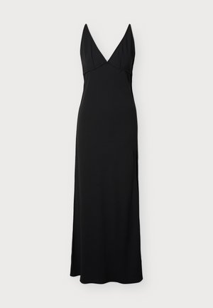 Mujer con un vestido largo negro sin mangas con un escote en V profundo y una abertura lateral, combinado con sandalias marrones de tacón con hebillas.