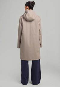 Beige vesimproof takki, jossa on huppu, pitkät hihat ja napit, yhdistettynä leveisiin tummansinisiin housuihin. Silkkinen pinta, yksinkertainen muotoilu.