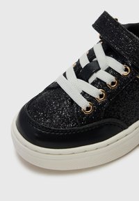 Sneaker glitter nera con lacci bianchi e occhielli dorati. Presenta una parte superiore testurizzata e una punta in pelle nera. Suola in gomma con motivo antiscivolo.