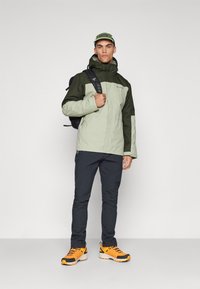 Veste verte bicolore résistant à l'eau avec une capuche, associée à un pantalon sombre et des chaussures orange et noires. Accessoire : sac à dos noir.