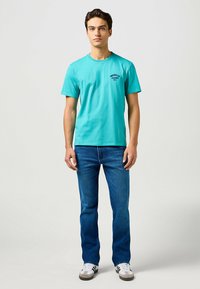 Turquoise katoenen T-shirt met ronde hals en borstlogo, gecombineerd met blauwe denim jeans en witte sneakers met grijze accenten.