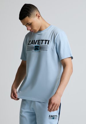 Jeune homme regardant vers le bas, portant un T-shirt Zavetti bleu clair et un short assorti avec un logo noir et bleu sur fond uni.