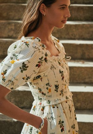 Naine kannab valget lillelise mustriga kleiti täispuhutavate varrukate ja volditud rinnaosaga, seisab külgvaates kiviastmetel pehme loomuliku valgusega.