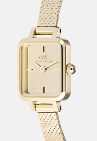 Montre rectangulaire en or avec un bracelet en mesh, cadran minimaliste présentant de fins repères horaires et une couronne sur le côté. Fini métallique lisse.