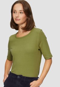 Prążkowany, krótki top w oliwkowej zieleni z bawełny. Posiada okrągły dekolt i dopasowany krój. Połączony z ciemnymi, teksturowanymi spodniami.