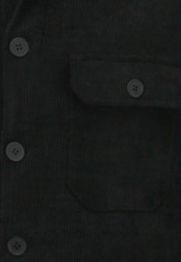 Zwart corduroy overhemd met vier zwarte knopen aan de voorkant en een borstzak met een omgevouwen klep. Getextureerde stof en effen kleur.
