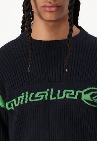 Pulover tricotat navy cu textură ribbed, guler rotund și un logo verde îndrăzneț pe piept.