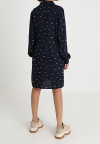 Abito camicia blu navy con maniche lunghe e colletto, decorato con un motivo sparso di piccoli disegni bianchi e dorati. Indossato con sneakers platform beige.