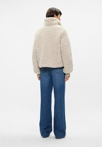 Beež faux fur jakk kõrge kaelusega, ümarate õlgade ja pikkade varrukatega, koos keskmise tõusuga siniste teksadega, millel on laiad jalad.