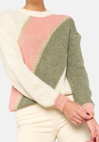 Kvinna i en stickad tröja med breda diagonala färgblock i kräm, rosa och olivgrön, med guldfärgad kant och ribbade ärmslut.