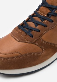 Brown leather and suede sneaker featuring navy laces, a cushioned white midsole, and detailed stitching along the upper.