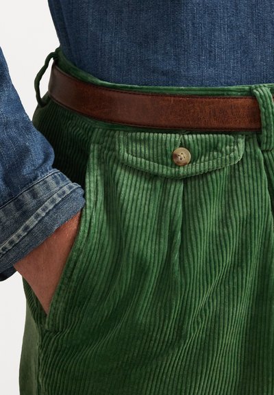 Green corduroy trousers with vertical ribs, featuring a brown leather belt and a buttoned pocket. Denim shirt cuffs are visible.