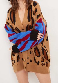 Leopardmönstrad cardigan med brun botten, kontrasterande ärmar i blå och röda ränder, samt svarta manschetter. Har knappstängning och stickad textur.
