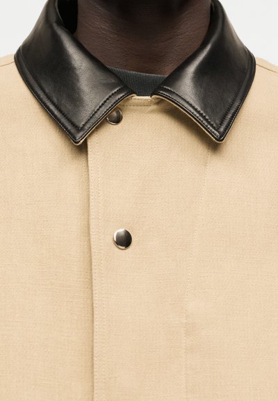 Μπεζ παλτό με λείο μαύρο δερμάτινο γιακά και δύο ορατά μεταλλικά κουμπιά, που φοριέται πάνω από σκουρόχρωμο ένδυμα.