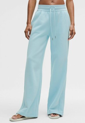 Lichtblauwe wijdvallende joggingbroek met een trekkoord in de taille, gedragen door een staande persoon in combinatie met witte slides.