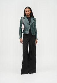 Zelena motoristična usnjena jakna s srebrno opremo, oblečena čez črno majico, združena s širokimi črnimi hlačami z obstranskimi žepi.