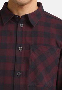 Chemise à carreaux rouge et noire, avec une poche poitrine, texture lisse et boutons noirs contrastés.