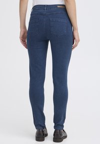 Jean en denim de lavage foncé avec un design ajusté, poches arrière et taille haute, associés à des chaussures noires. Texture lisse et style classique.