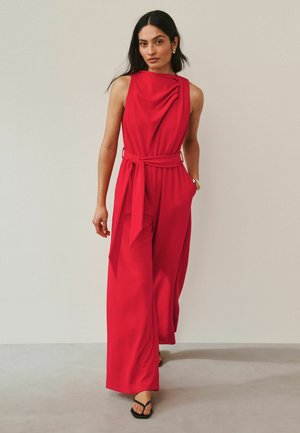 Frau geht nach vorne, trägt einen ärmellosen roten Jumpsuit mit drapiertem Ausschnitt und Taillengürtel, kombiniert mit schwarzen Sandalen mit offenen Zehen.