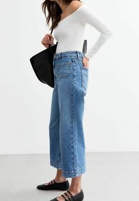 Vaqueros de tiro alto azul claro con corte de pierna ancha, combinados con un top blanco sin hombros y zapatos planos negros. Se incluye un bolso tote negro.