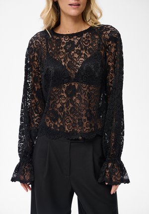 Frau in schwarzer, floral gemusterter Spitzenbluse mit langen Ärmeln und Rüschenelementen, kombiniert mit schwarzen Hosen, die ein transparentes, strukturiertes Muster zeigen.