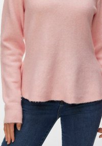 Femme portant un pull en maille rose clair à manches longues et un jean bleu foncé, debout avec les bras détendus le long du corps.