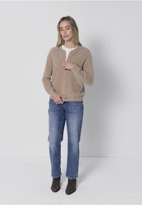 Cardigan beige à fermeture éclair en texture douce, porté sur une chemise blanche. Associ é à un jean bleu et des bottines marron, debout devant un fond clair.